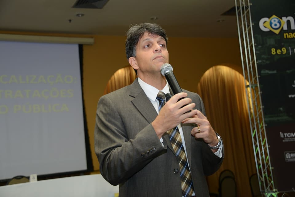 Palestrante de terno fala ao microfone diante do telão, com banner do evento ao fundo.