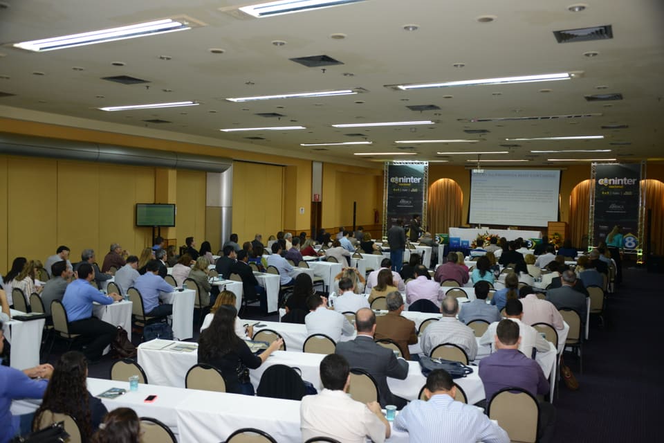 Sala de eventos cheia, com participantes sentados em mesas retangulares voltadas para o telão de uma apresentação.
