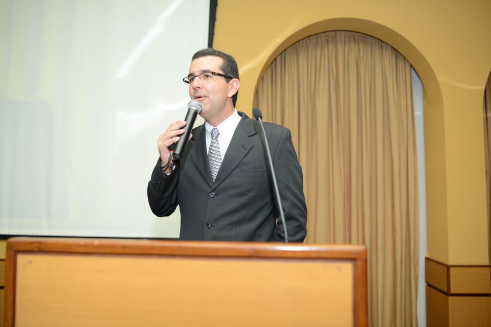 Homem de terno fala ao microfone atrás de uma tribuna de madeira, diante de uma tela de projeção.