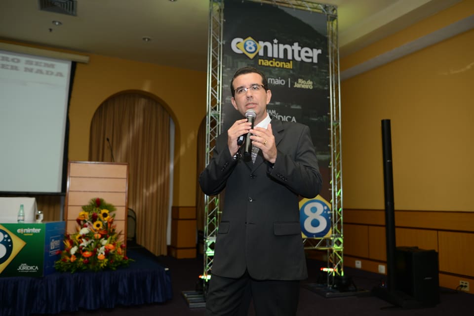 Homem de terno fala ao microfone diante do painel vertical do evento, com arranjo de flores e tribuna ao lado.