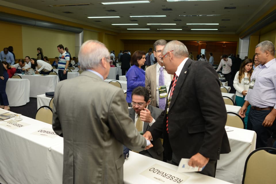 Dois homens se cumprimentam com aperto de mão sobre a mesa durante o intervalo, cercados por outros participantes.