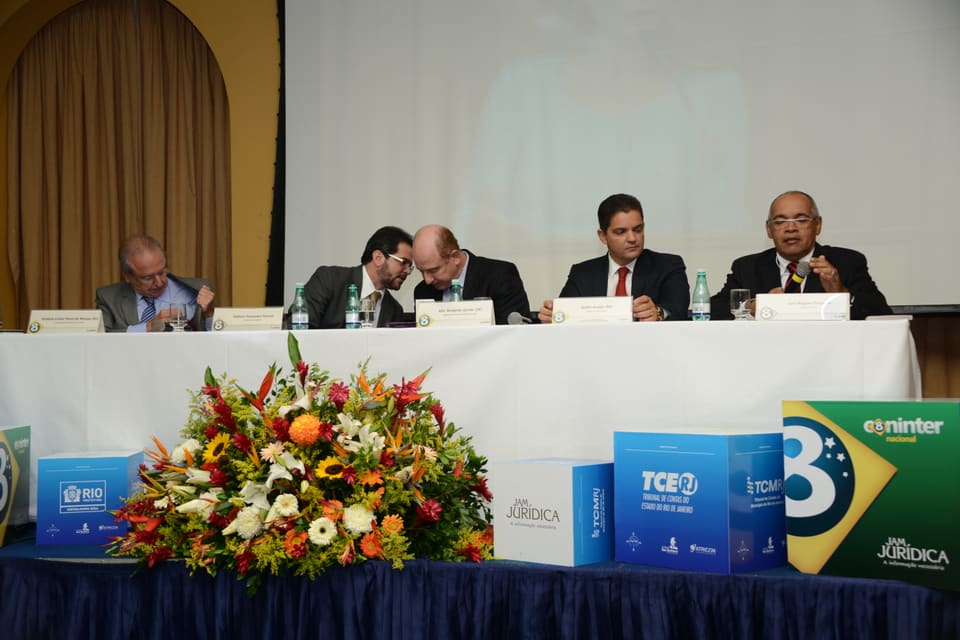 Mesa de abertura com autoridades sentadas, arranjo de flores e cubos com marcas de tribunais de contas.