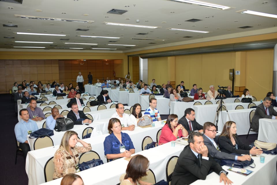 Plateia de profissionais sentados em mesas compridas acompanha uma apresentação em sala de eventos.