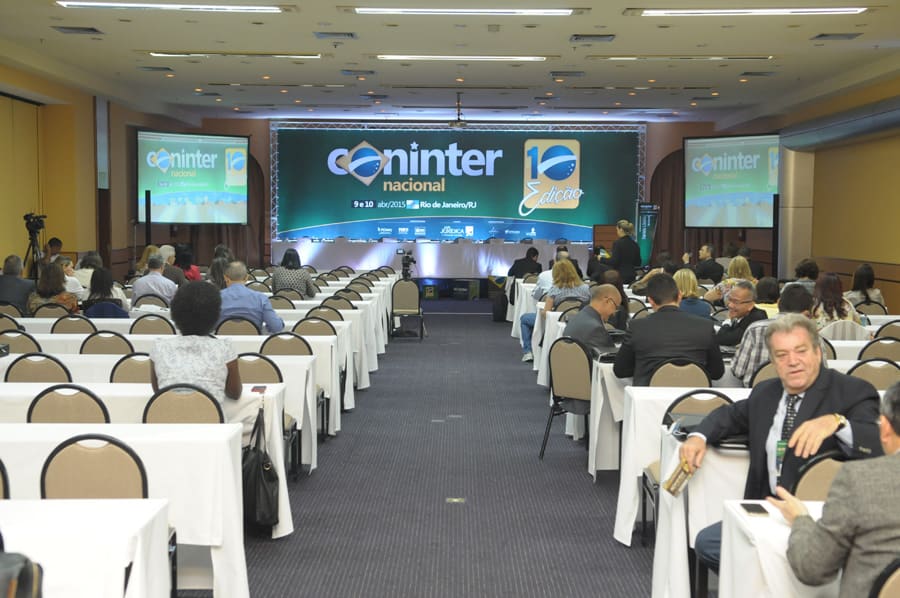 Sala de eventos vista do fundo, com mesas alinhadas e o telão principal exibindo a marca do congresso.