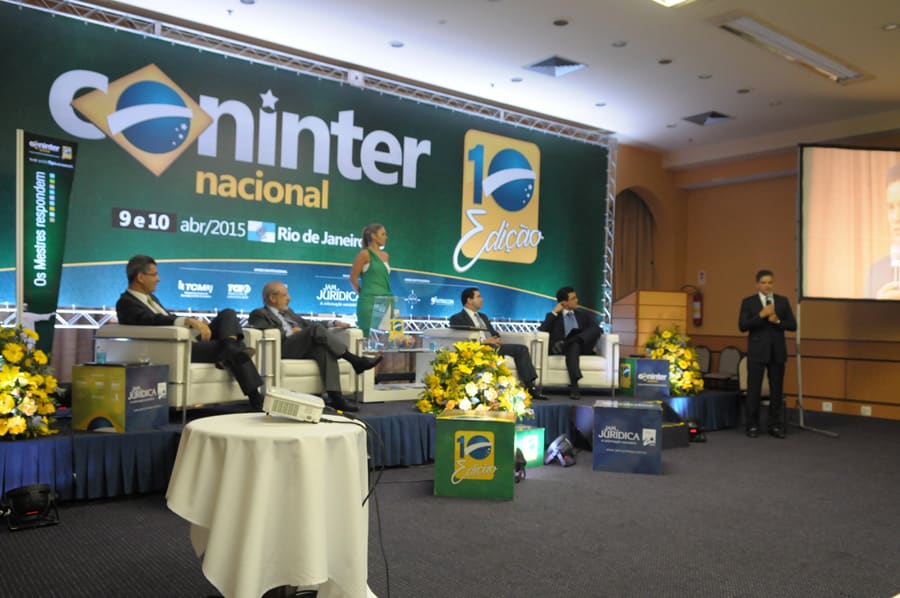 Painel de debate com quatro convidados sentados em poltronas no palco e um mediador em pé ao microfone.