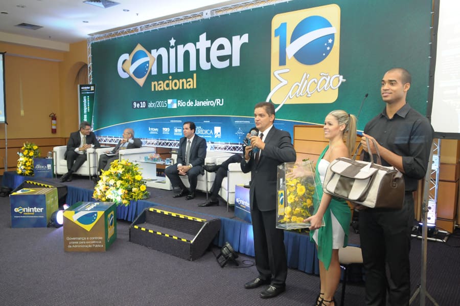Apresentador fala ao microfone no palco, com os convidados do painel sentados atrás.